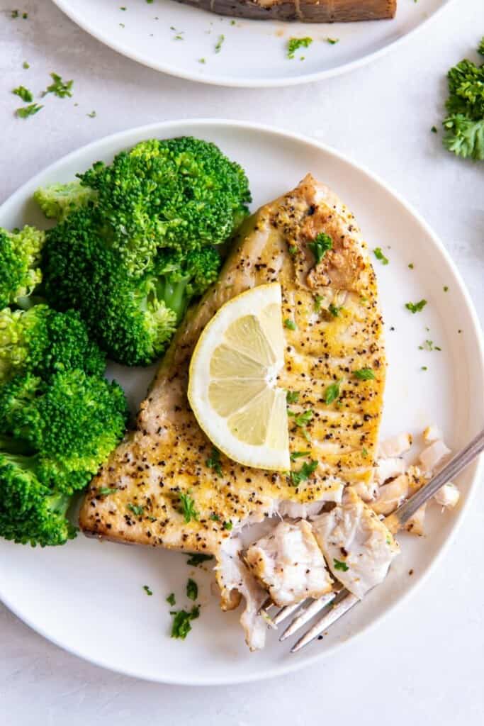 Air Fryer Swordfish on a white plate topped with lemon, served with broccoli.