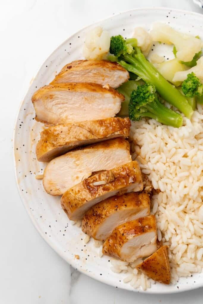 Overhead view of sliced chicken breast with rice and broccoli on a white plate.