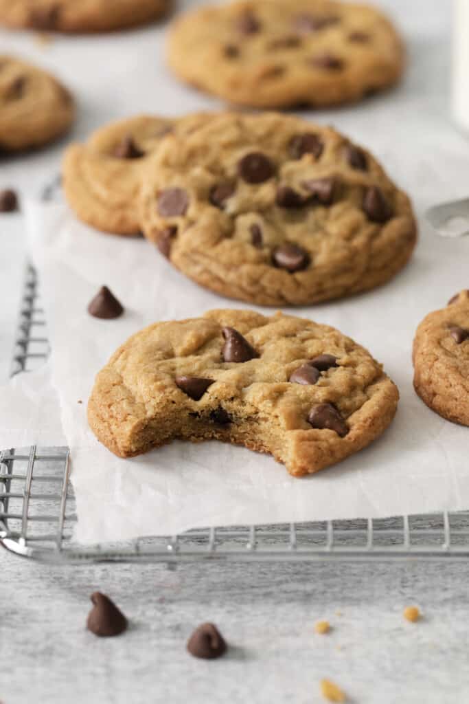 Baked chocolate chip cookies on a cooling rack lined with parchment paper. One cookie has a bite taken out of it.