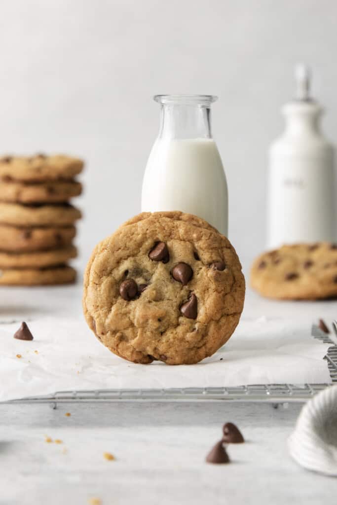 Chocolate Chip Cookie lifted up and resting against a bottle of milk. Additional cookies and bottle of milk in background.