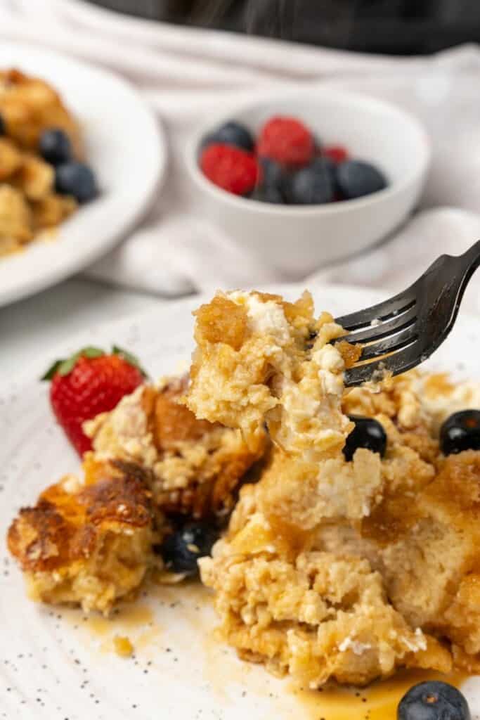 A fork holding a bite of the single serving of crockpot french toast casserole on a white plate with berries in the background.