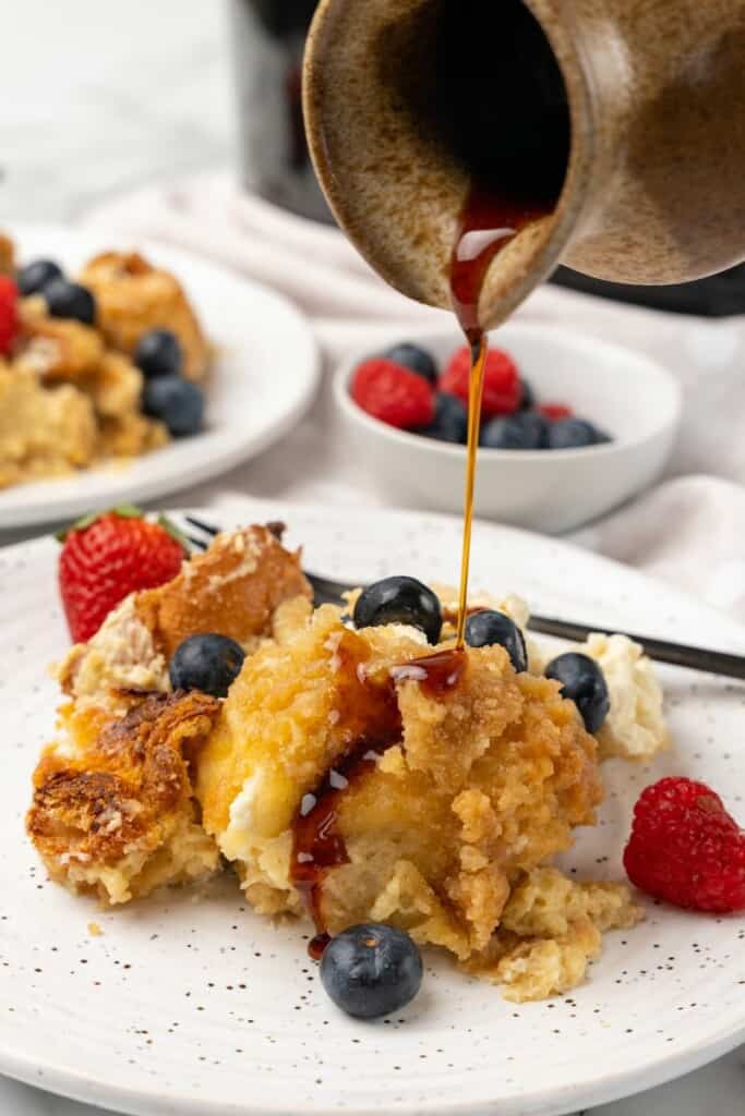 Single serving of crockpot french toast casserole with berries on a white plate with syrup being poured over the top.