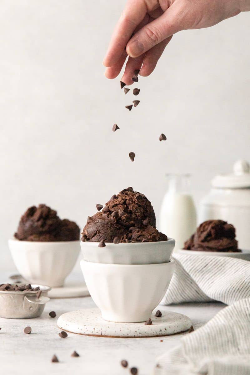 Scoops of edible brownie batter in white bowls on saucers. A hand overhead dropping chocolate chips on top of the brownie batter.