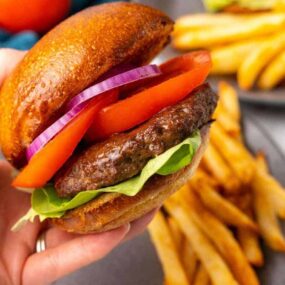 Closeup view of a fully dressed hamburger that has been prepared in a Ninja Foodi with a gray plate holding french fries in the background.