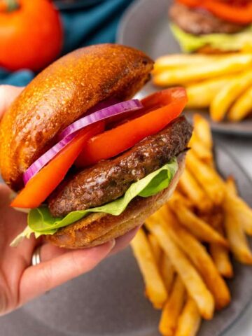 Closeup view of a fully dressed hamburger that has been prepared in a Ninja Foodi with a gray plate holding french fries in the background.