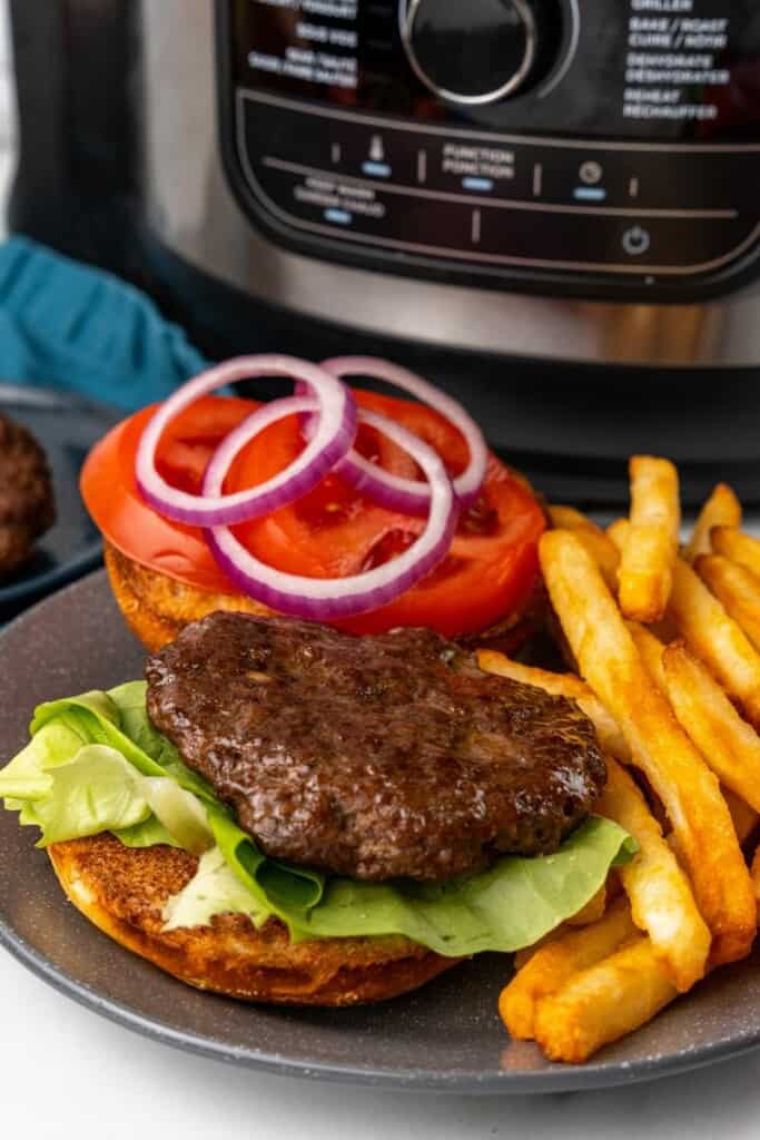 A hamburger patty on a bun with lettuce, tomato, pickles, and onion next to french fries on a gray plate with a Ninja Foodi in the background.