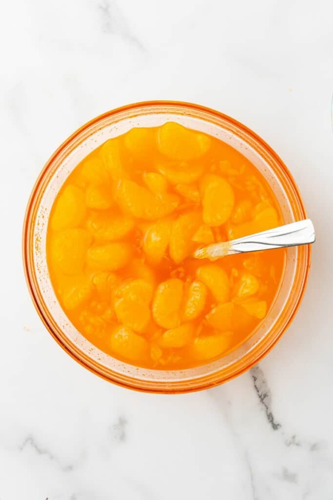 Overhead view of liquid Jello mix with mandarin oranges in a clear bowl with a spoon.