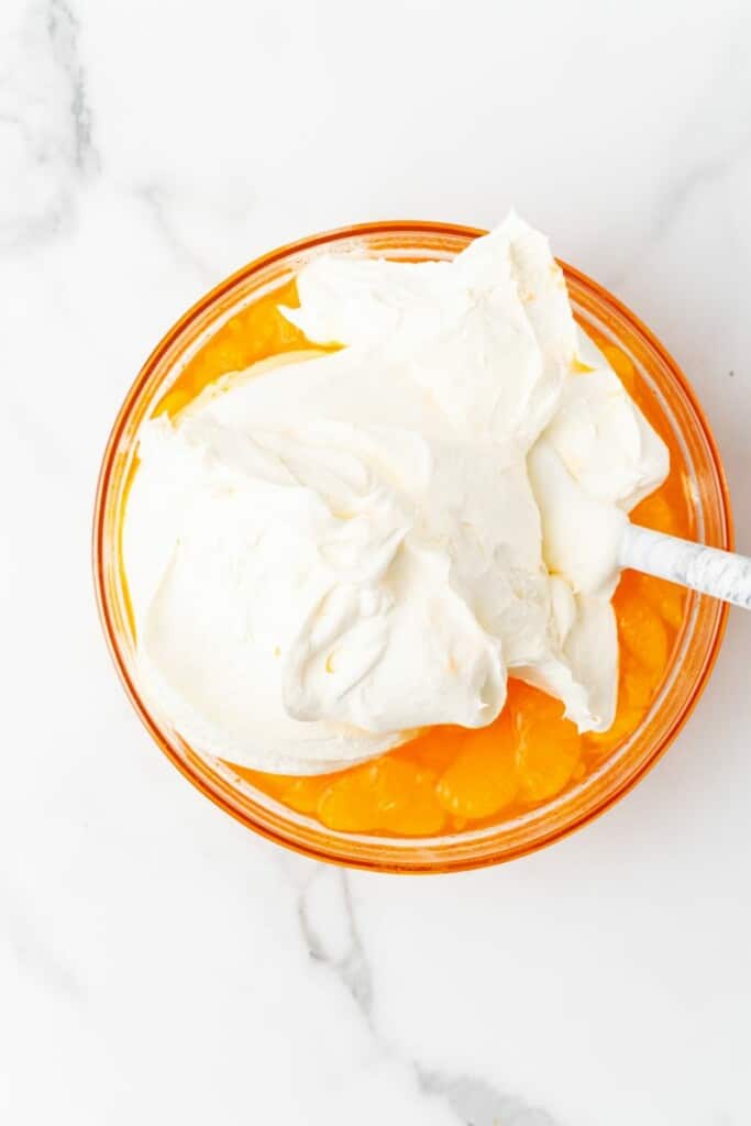 Overhead view of cool whip on top of Jello & Mandarin orange mix in a clear bowl with a spoon.