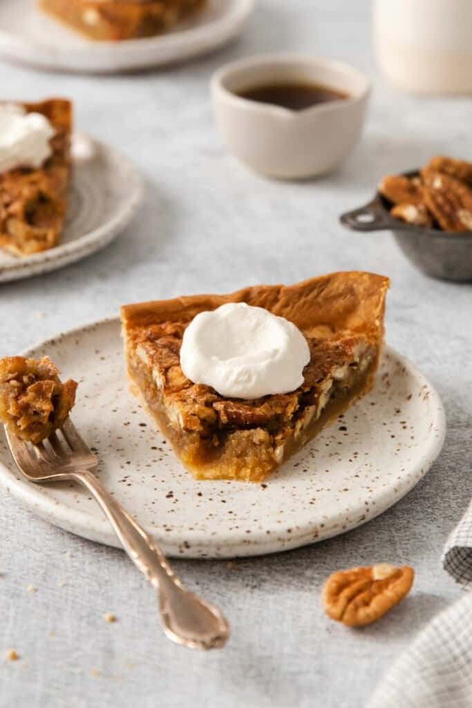Slice of pecan pie topped with whipped cream. One bite removed resting on a fork all on an off white plate.