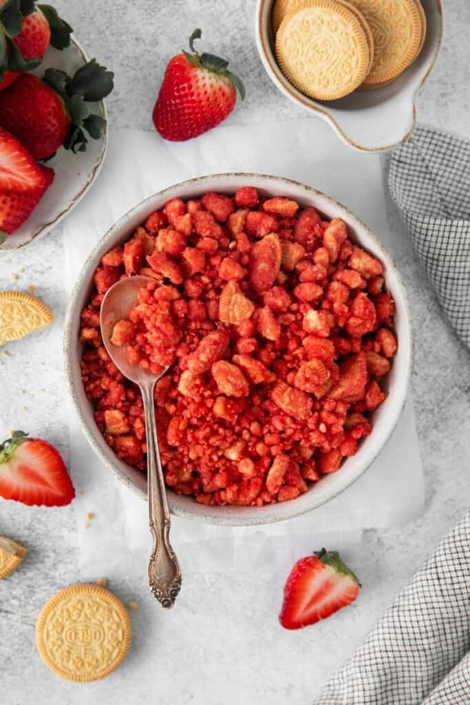 Overhead view of strawberry crunch in a white bowl with a spoon. Oreos and strawberries in background.