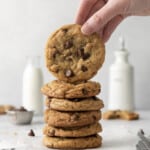 A pile of baked chocolate chip cookies with one raised, bottles of milk in the background.