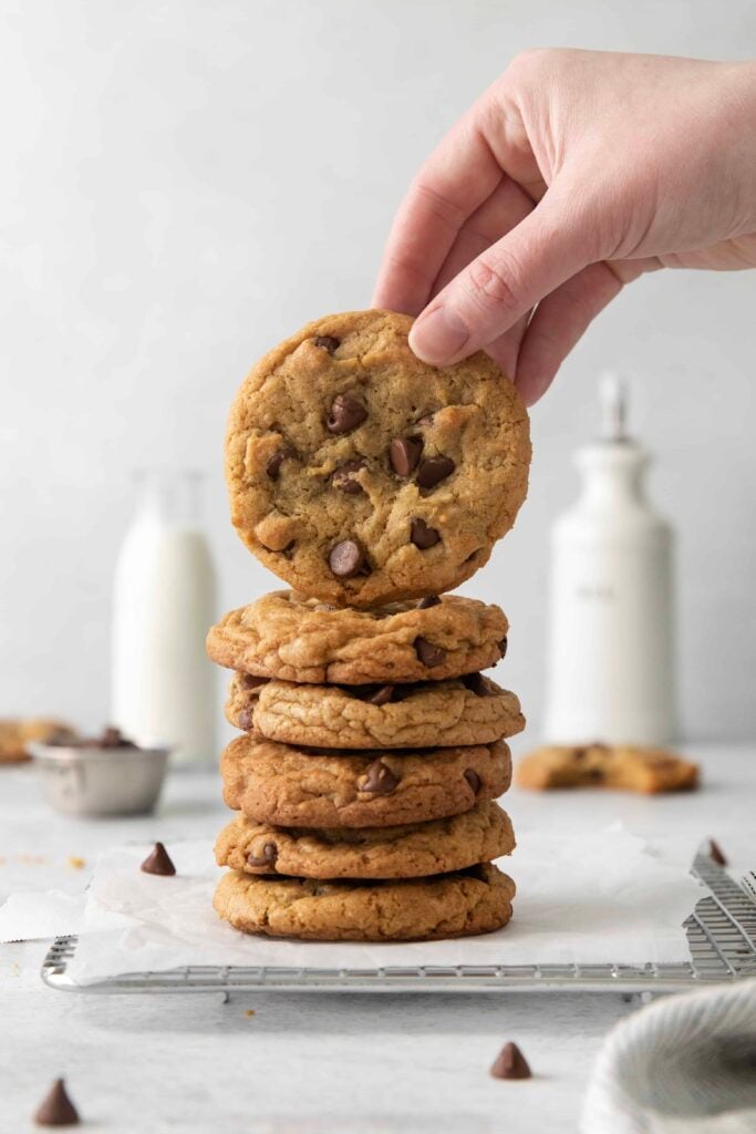 Stack of baked chocolate chips cookies with one lifted, bottles of milk in the background.