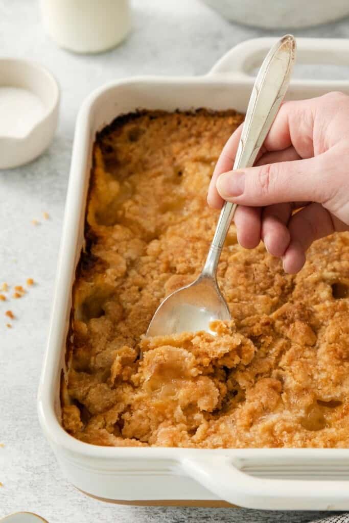 A spoon lifting a bite of peach cobbler out of a square white baking dish.