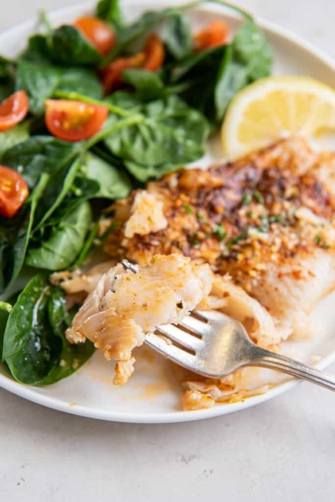 A fork lifting a bite of air fryer flounder from a white plate.