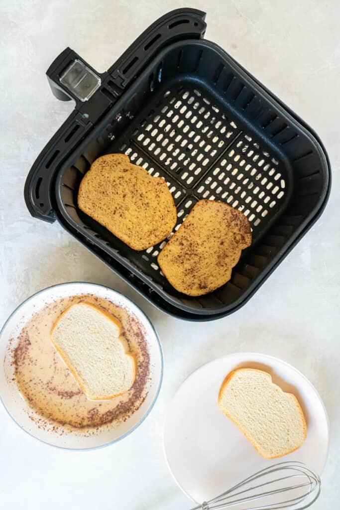 Two slices of french bread in a black air fryer basket. Bowls of cinnamon sugar and liquid ingredients alongside.
