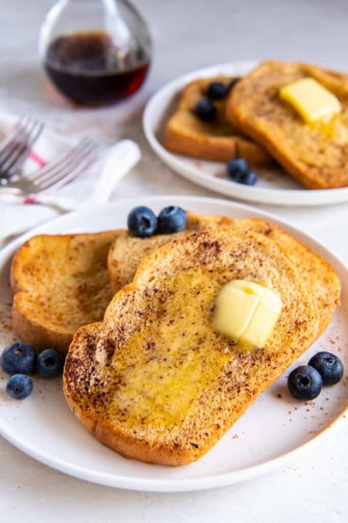 Slices of french toast on white plates with butter and blueberries.