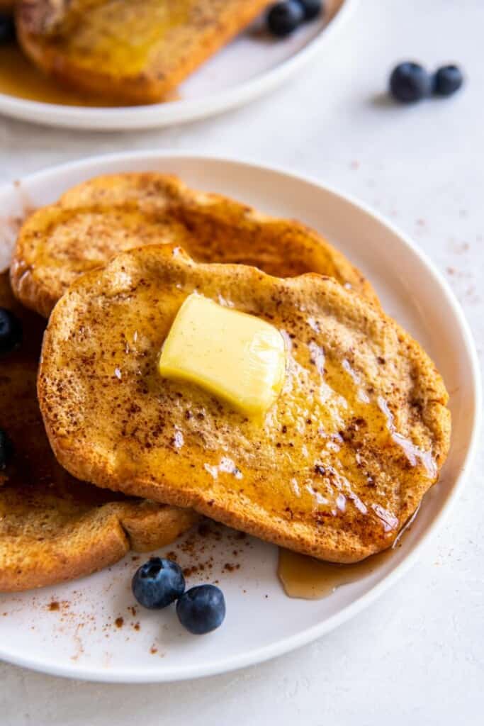 Slices of french toast on a white plate with blueberries and butter.