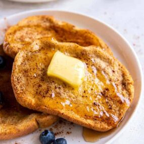 Slices of french toast on a white plate with butter & blueberries.