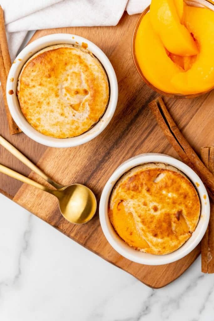 Two peach cobblers in white ramekins resting on a wooden board with peaches in the background.