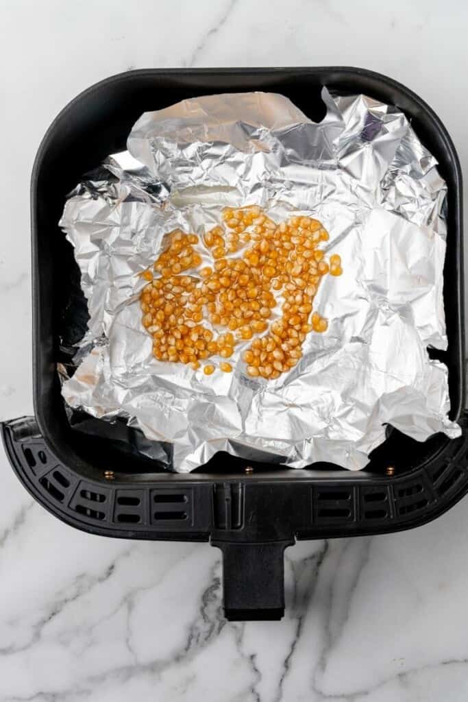Popcorn kernels spread in a single layer on tin foil inside an air fryer basket.