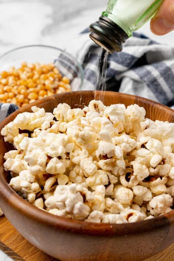 Adding a dash of salt to popcorn in a wooden bowl with kernels in the background.