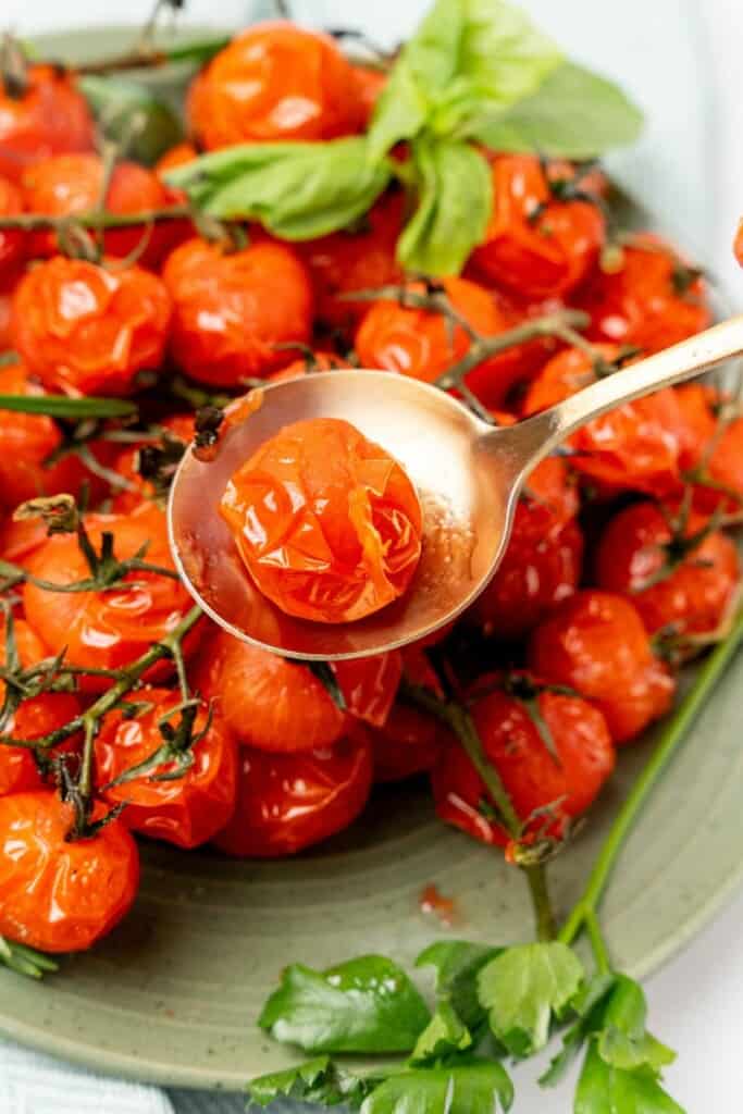 Closeup view of a spoon holding a single air fried cherry tomato above a plate of additional cherry tomatoes.