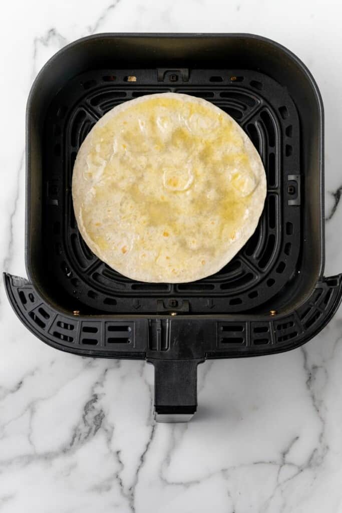 Tortilla spread with oil in the bottom of a black air fryer basket.