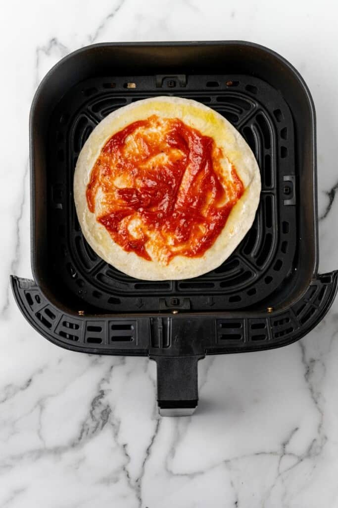 Sauce spread on a tortilla in a black air fryer basket.