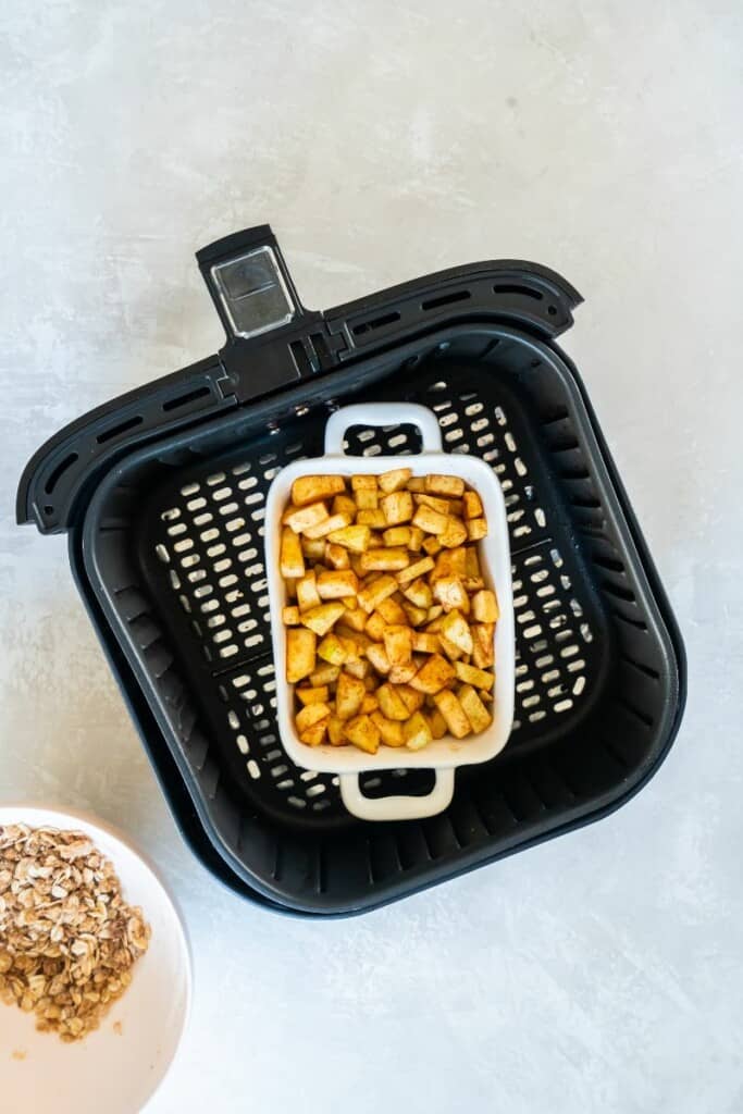 Apple crisp in a white dish inside a black air fryer basket waiting for prepared topping.