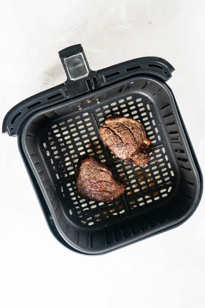 Two filet mignon steaks that have been air fried, resting in a black air fryer basket.