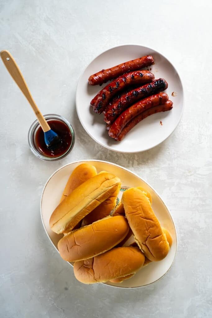 Grilled BBQ Hot Dogs on a white plate next to a plate of buns and a small bowl of bbq sauce.