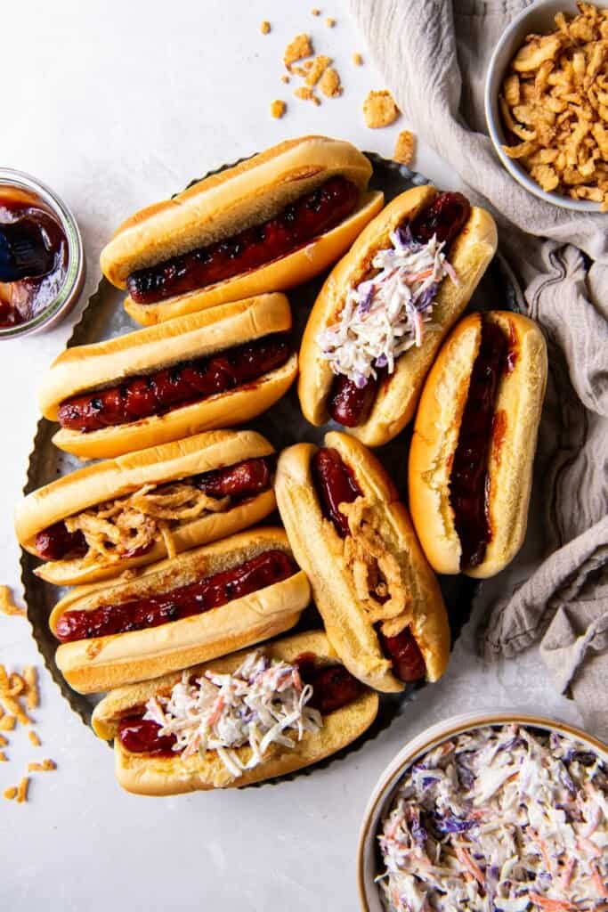 Overhead view of eight grilled bbq hot dogs on a plate with buns and toppings.