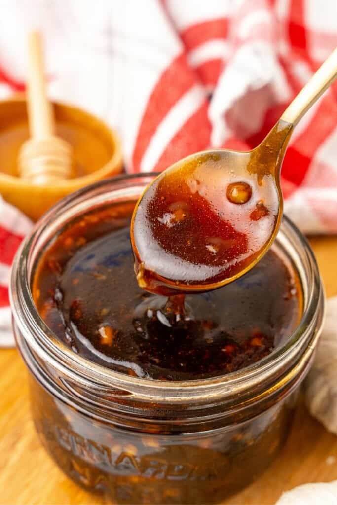 A spoon lifting a small amount of Honey Garlic Sauce out of a small clear mason jar.