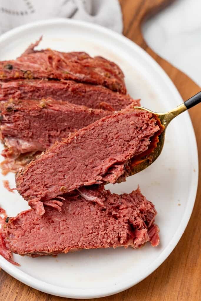 A serving fork holding a slice of corned beef brisket. Other slices resting on a white plate.