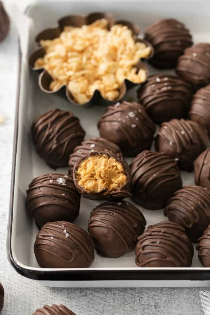 A pan with chocolate dipped peanut butter balls with rice krispies. One has a bite taken out to reveal the center.