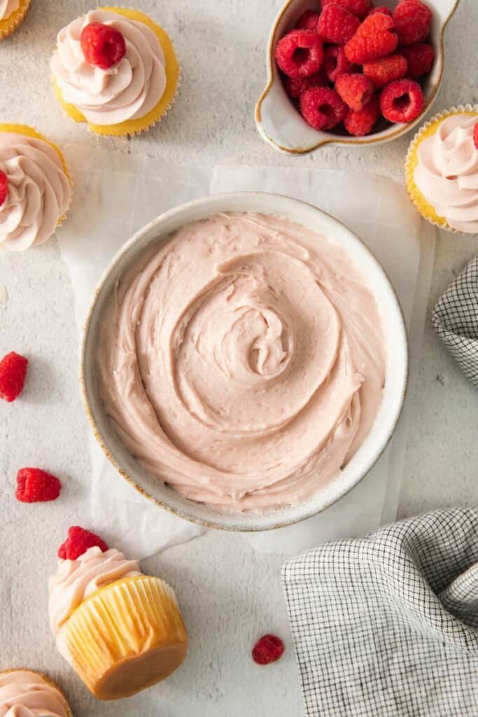 A bowl of raspberry jam cream cheese frosting surrounded by cupcakes and raspberries.