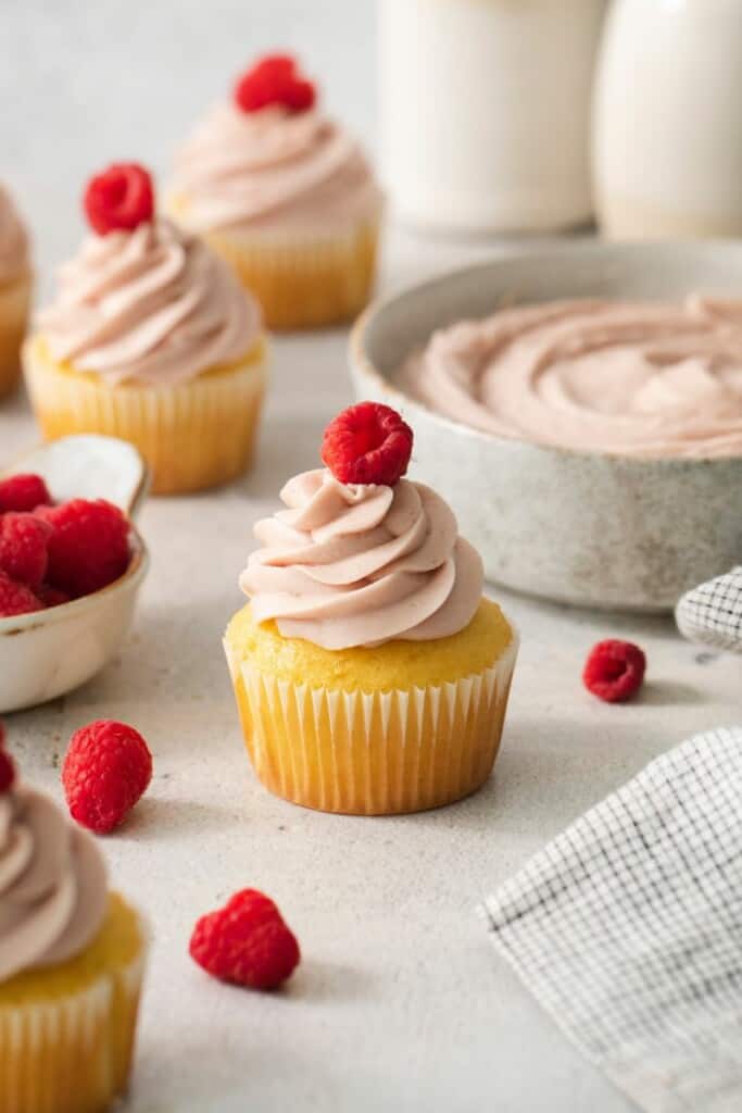 Several cupcakes topped with fresh raspberry jam cream cheese frosting and fresh raspberries.