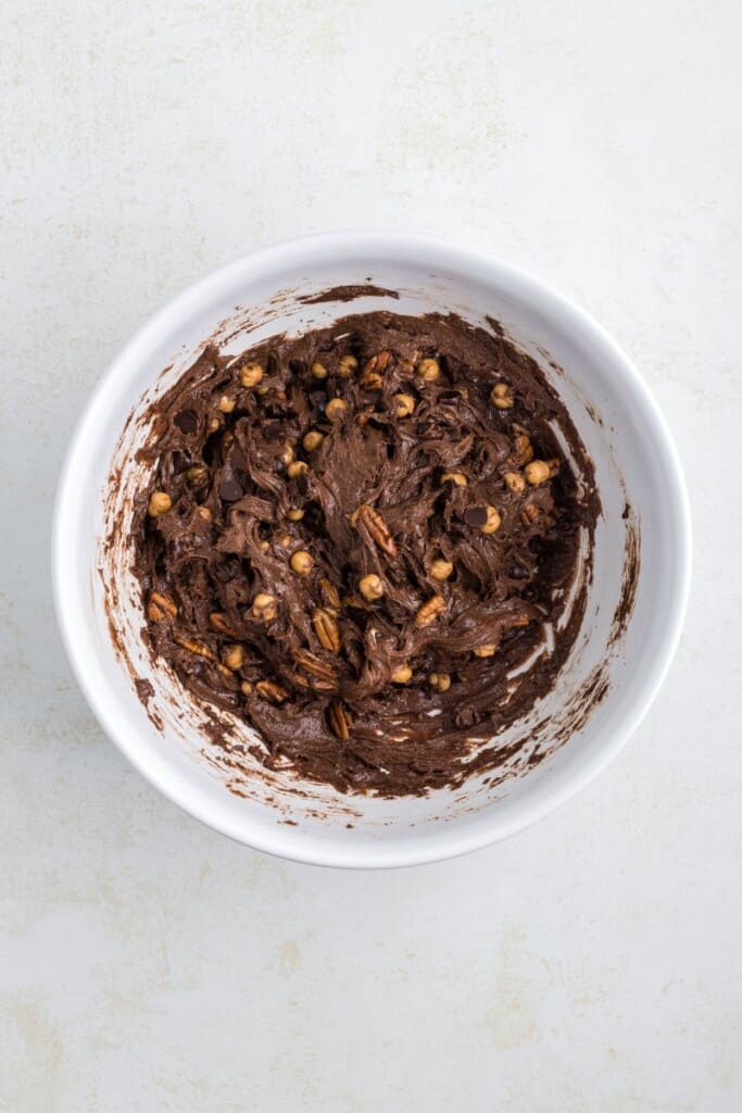 Folding in toasted pecans, caramel bits and chocolate chips with the cookie batter in a white mixing bowl.