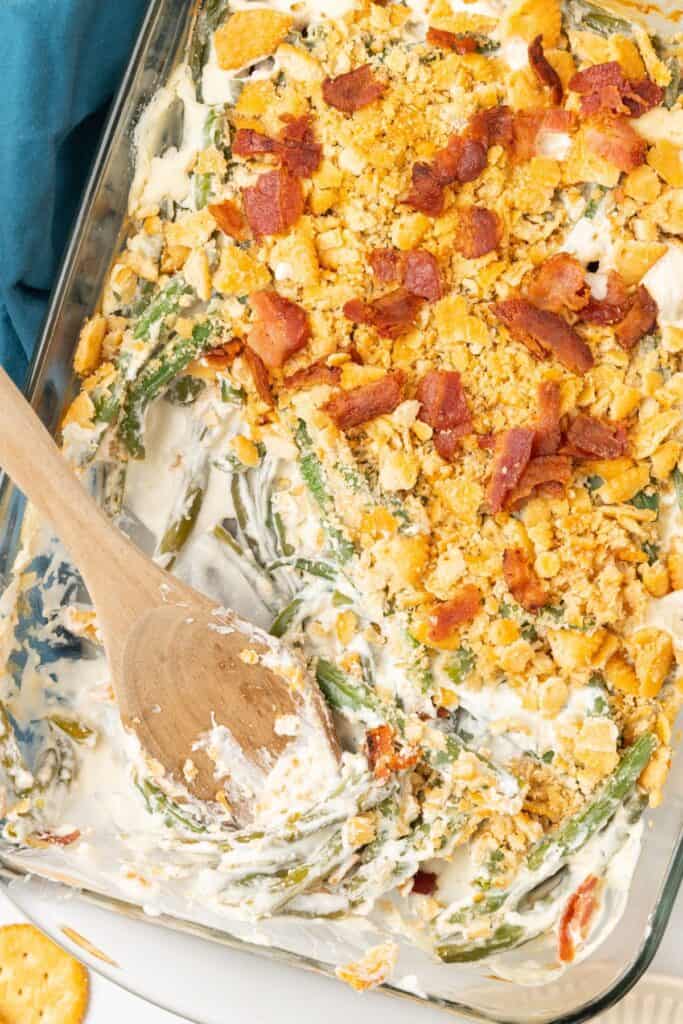 Green bean casserole with cream cheese in a casserole dish with some already scooped out and a wooden spoon in it.