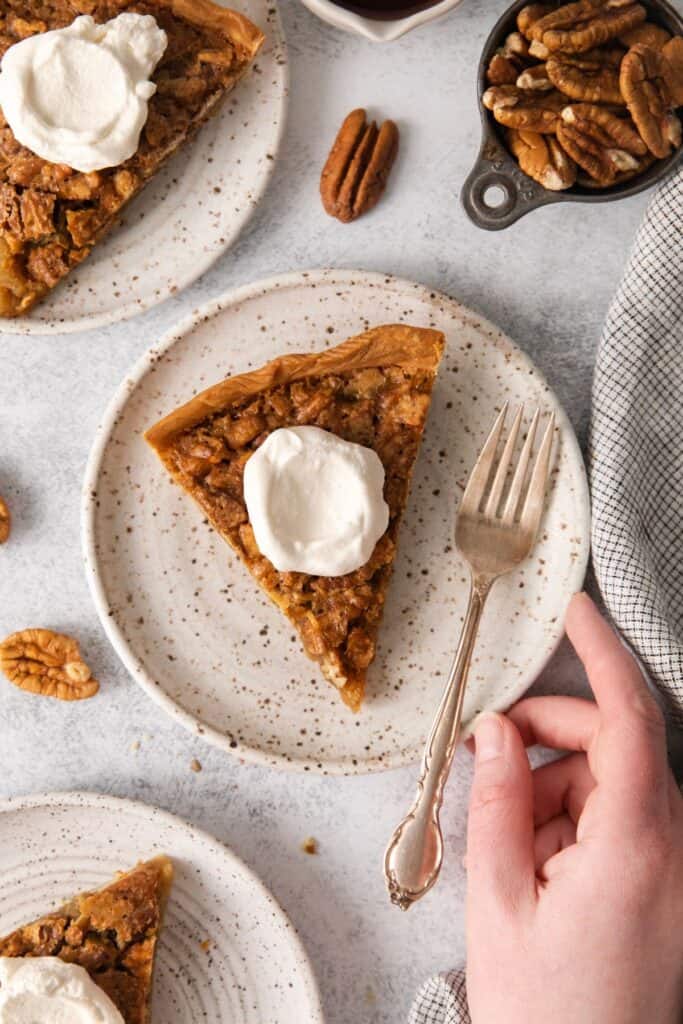 Pecan pie without corn syrup slice with whipped cream on top on a plate with a fork and a hand grabbing the plate.