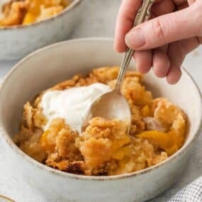 4 ingredient peach cobbler in a bowl with whipped cream with a spoon in it.