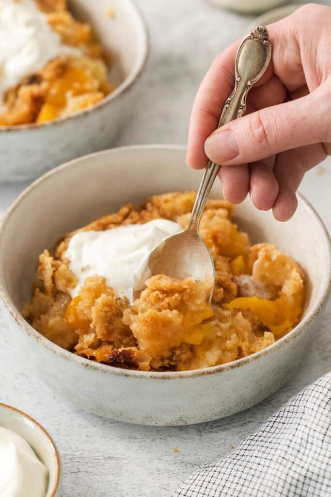 4 ingredient peach cobbler in a bowl with whipped cream with a spoon in it.