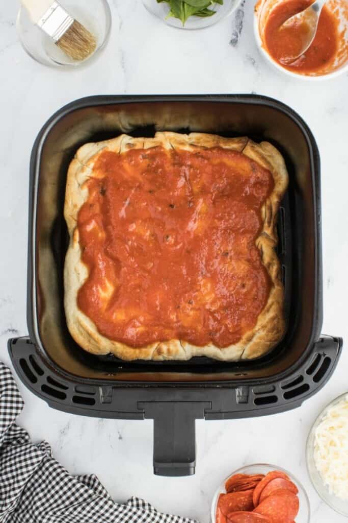 Pizza sauce spread over a baked pizza dough in a black air fryer basket.