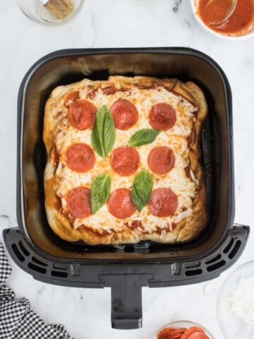 A pepperoni pizza baked in the air fryer, topped with basil, resting in a black air fryer basket.