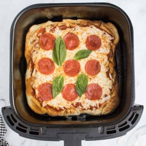 A pepperoni pizza baked in the air fryer, topped with basil, resting in a black air fryer basket.