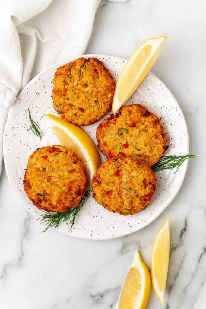Four prepared salmon patties on a speckled plate with lemon wedges.