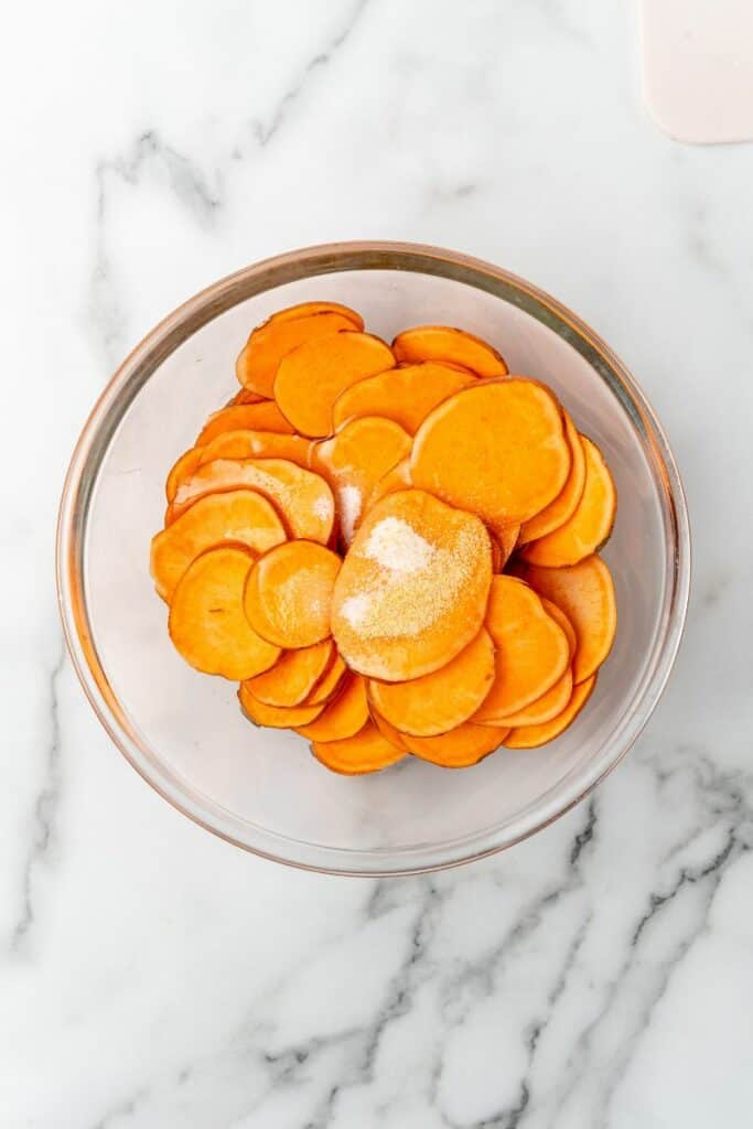 Slices of sweet potatoes in a clear glass bowl with seasoning and spices.