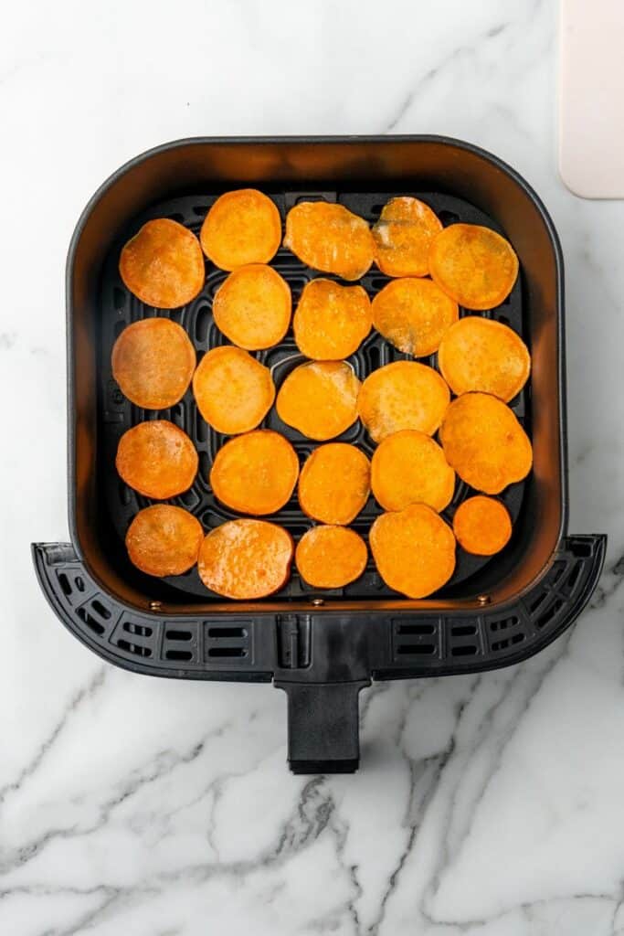Slices of sweet potatoes in an even layer resting in a black air fryer basket.