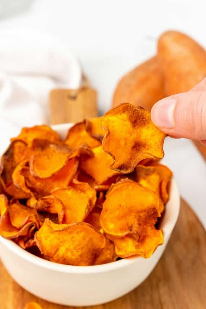 A hand lifting a sweet potato chip out of a white bowl.