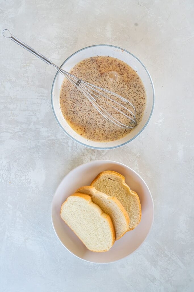 Air fried french toast batter whisked in a bowl with bread on a plate underneath it ready to be dipped.